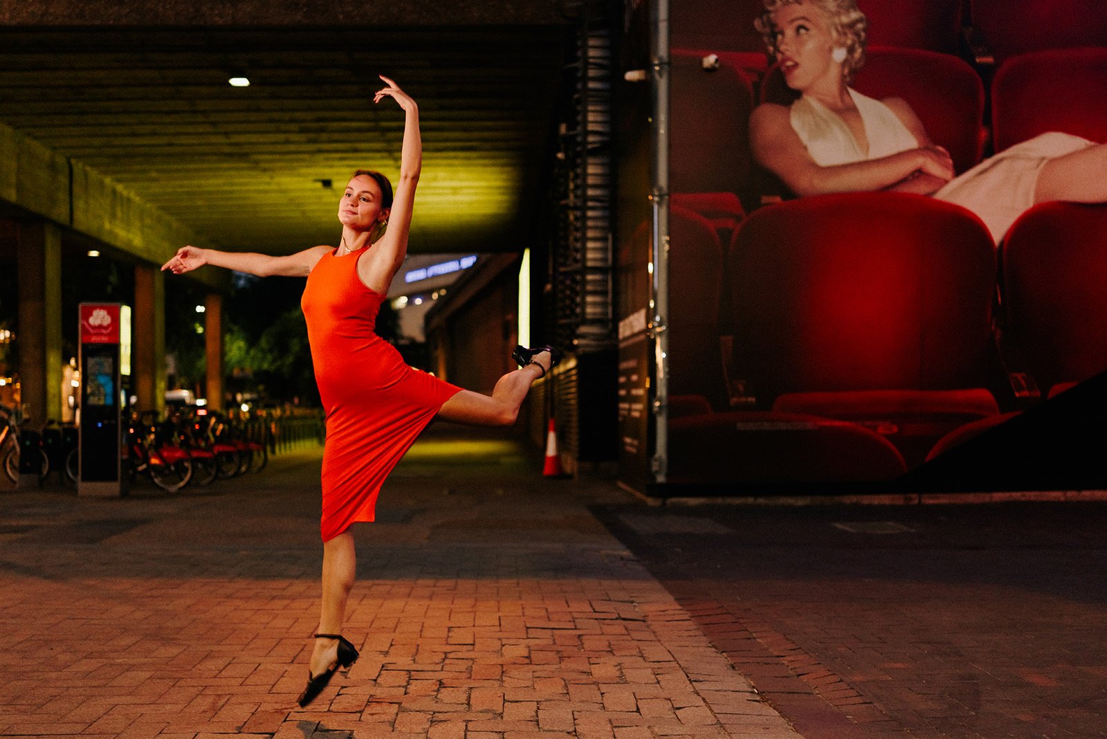 A ballet dancer in red dress dances at dusk in London south bank next to a bridge and a billboard of Marilyn Monroe.